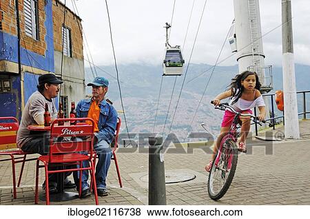 Men sitting in a street café, girl riding a bicycle, Metrocable cable car at back, slums, Comuna 13, Medellin, Colombia, South America, Latin America, America, South America View Large Photo Image Stock Photo - Men sitting in a street café, girl riding a bicycle, Metrocable cable car at back, slums, Comuna 13, Medellin, Colombia, South America, Latin America, America, South America. Fotosearch