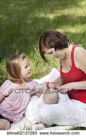 Stock Image - Mother giving her baby a bottle while a young girl watches. Fotosearch