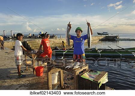 Stock Image - Papuan fishmonger holding two fish in the air on display before gutting them, beside him are children who will also gut customers fish for a fee, at the fish market in Kota Biak, Biak Island, West Papua, Indonesia, Southeast Asia, Asia. Fotosearch