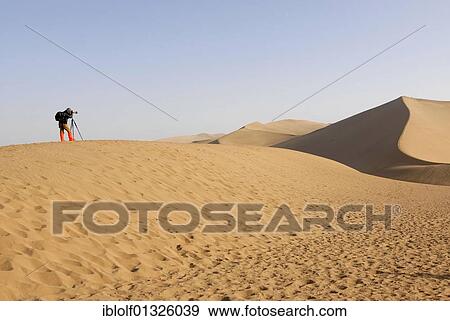 Photographer using a tripod and wearing red boots while standing on a sand hill in front of the sand dunes of the Gobi Desert and Mount Mingshan near Dunhuang, Silk Road, Gansu, China, Asia View Large Photo Image Stock Photo - Photographer using a tripod and wearing red boots while standing on a sand hill in front of the sand dunes of the Gobi Desert and Mount Mingshan near Dunhuang, Silk Road, Gansu, China, Asia. Fotosearch