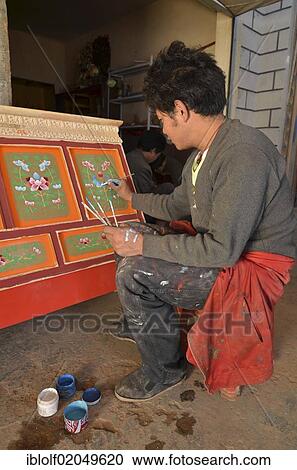 Stock Image - Tibetan handicrafts, carpenter painting a typical Tibetan wooden cabinet with red paint and ornaments, lacquer work, Tibet, China, Asia. Fotosearch