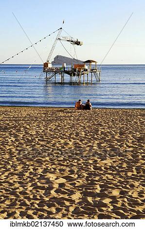 Tourist couple sitting on Playa Levante beach in front of a water ski facility, Benidorm, Spain, Europe View Large Photo Image Stock Image - Tourist couple sitting on Playa Levante beach in front of a water ski facility, Benidorm, Spain, Europe. Fotosearch
