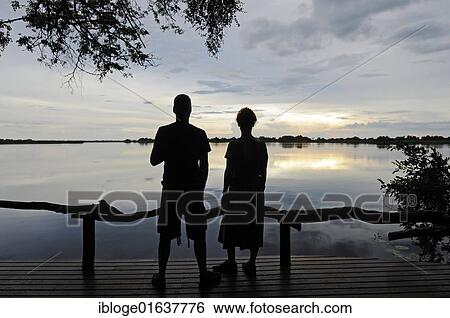 Stock Photograph - Tourists sitting on the terrace of a lodge in the evening, Guma Lagoon, Guma Lagoon, Okavango Delta, Botswana, Africa. Fotosearch