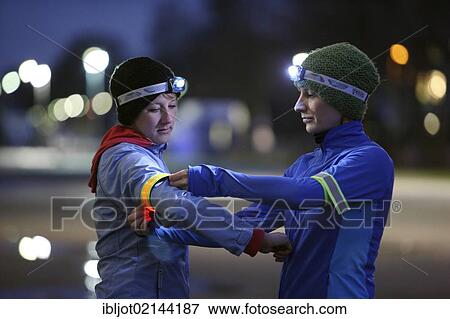 Two young women preparing to go jogging at dusk in winter, putting on reflector strips and LED headlamps, wearing reflective wind and waterproof performance apparel View Large Photo Image Stock Photo - Two young women preparing to go jogging at dusk in winter, putting on reflector strips and LED headlamps, wearing reflective wind and waterproof performance apparel. Fotosearch