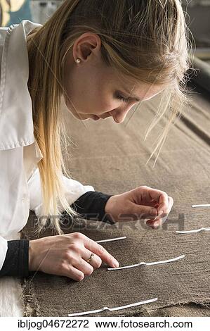 Stock Image - Restoration studio, restorer, woman sews finished support fabric on lent scarf at paper strip marking, Munich, Bavaria, Germany, Europe. Fotosearch