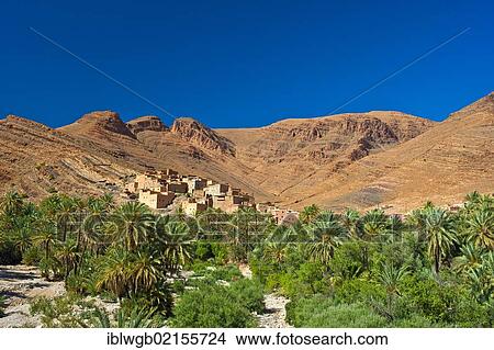 Picture - Small clay village and a palm grove in front of the red mountain landscape in the Ait Mansour valley, Anti-Atlas mountains, southern Morocco, Morocco, Africa. Fotosearch