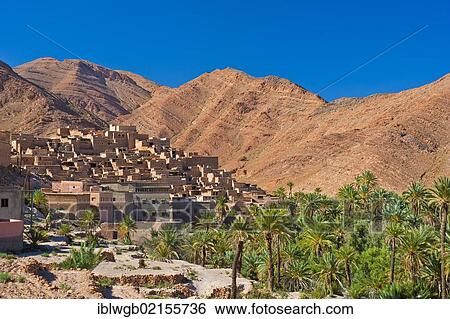 Stock Photograph - Small clay village and a palm grove in front of the red mountain landscape in the Ait Mansour valley, Anti-Atlas mountains, southern Morocco, Morocco, Africa. Fotosearch
