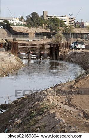 Picture - The heavily-polluted New River, as it enters the USA from Mexico, the pollution results from a mix of untreated sewage, agricultural runoff, and industrial waste; a U. S. Border Patrol agent is parked on the right bank near the border fence, the.... Fotosearch