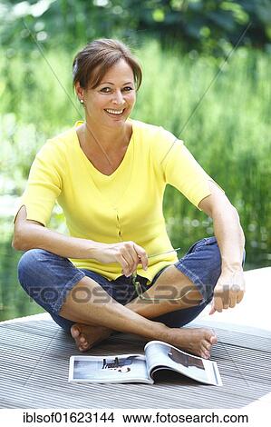 Picture - Woman, 45 years, with glasses and a magazine, wearing casual clothes, sitting on a wooden footbridge. Fotosearch