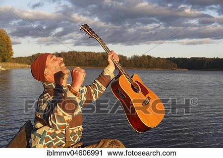 Stock Image - Man plays guitar on a boat landing stage at the lake and is pleased, Mecklenburg Lake District, Mecklenburg-Western Pomerania, Germany, Europe. Fotosearch