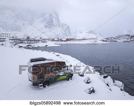 Stock Photo - Motorhome painted as an alpine hut in snowfall at the parking lot in front of Reinefjord, behind mountains in fog and fishing buildings, Reine, Nordland, Lofoten, Norway, Europe. Fotosearch