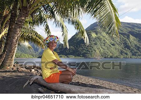 Stock Image - Old woman with wreath of flowers sitting under palm trees on tree trunk, coast near Taurita, Tahiti Iti, society islands, Windward Islands, French Polynesia, Oceania. Fotosearch