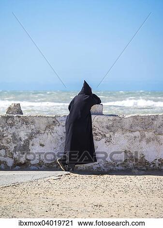 Stock Image - Ein Berber mit schwarzer Djellaba lehnt an der Hafenmauer, Place Moulay Hassan, Essaouira, Marokko, Afrika. Fotosearch