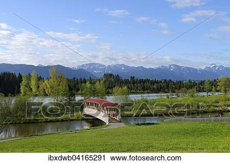 Stock Image - Gedeckte Brucke uber einen Seitenarm des Lech mit Ammergauer Alpen, bei Lechbruck, Bayerisch Schwaben, Bayern, Deutschland, Europa. Fotosearch