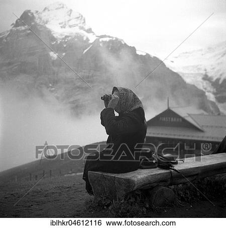 Stock Photograph - An elderly woman sits in the mountains on a wooden bench and examines the landscape through binoculars, ca. 1945 to 1955, Switzerland, Europe. Fotosearch