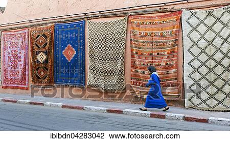 Stock Image - Moroccan woman walking past carpets hanging on wall, Marrakech, Morocco, Africa. Fotosearch