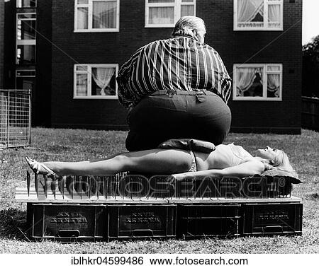 Stock Photograph - Thick man sitting on a woman lying on nail board, England, Great Britain. Fotosearch