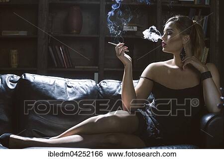 Stock Photograph - Young woman smoking a cigarette on a dark sofa. Fotosearch