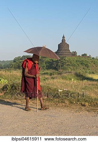Stock Image - Buddhist monk with umbrella walks on dirt-road in Mrauk U, Burma, Myanmar, Asia. Fotosearch