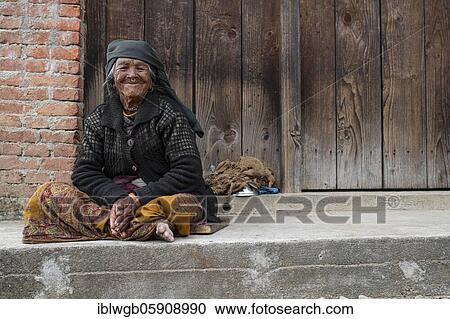 Stock Image - Friendly smiling elderly woman with headscarf and nose ring sitting in front of a wooden door, Himalaya, Jiri, Khumbu region, Nepal, Asia. Fotosearch