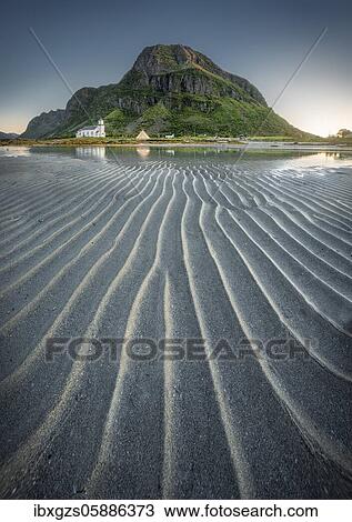 Stock Image - Gimsoy church reflected in the sea at low tide, in front sand beach with ribbed structure, behind mountain in evening sun, green vegetation on mountain slopes, Lofoten, Nordland, Norway, Europe. Fotosearch
