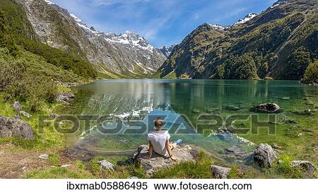 Stock Photography - Hiker sitting on the shore, Lake Marian, Fiordland National Park, Te Anau, Southland, South Island, New Zealand, Oceania. Fotosearch