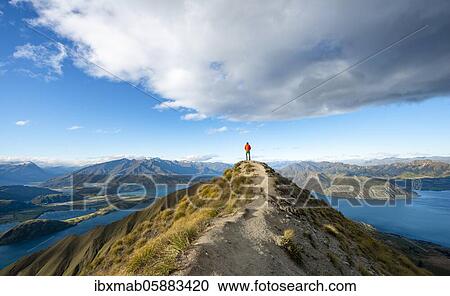 Stock Image - Hiker stands at the summit, view of mountains and lake from Mount Roy, Roys Peak, Lake Wanaka, Southern Alps, Otago, South Island, New Zealand, Oceania. Fotosearch