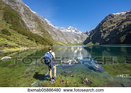 Stock Image - Hiker stands on shore, Lake Marian, Fiordland National Park, Te Anau, Southland, South Island, New Zealand, Oceania. Fotosearch