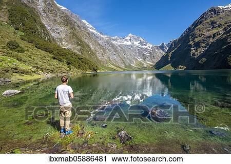 Stock Image - Hiker stands on shore, Lake Marian, Fiordland National Park, Te Anau, Southland, South Island, New Zealand, Oceania. Fotosearch