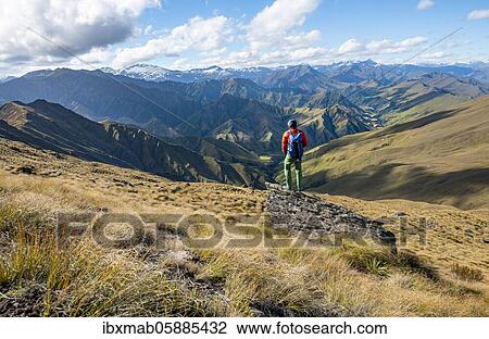 Stock Image - Hiker stands on stone, hiking trail to Ben Lomond, view of mountains, Southern Alps, Otago, South Island, New Zealand, Oceania. Fotosearch