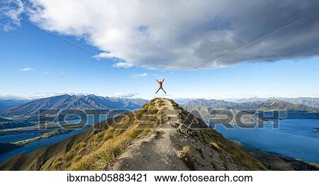 Stock Image - Hiker takes a skydive, views of mountains and lake from Mount Roy, Roys Peak, Lake Wanaka, Southern Alps, Otago, South Island, New Zealand, Oceania. Fotosearch