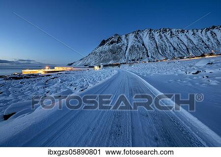 Stock Image - Iced snow road, illuminated village in twilight, residential and industrial buildings, Nordland, Lofoten, Norway, Europe. Fotosearch