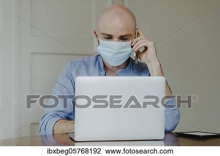 Stock Image - Man with face mask at home office. Fotosearch