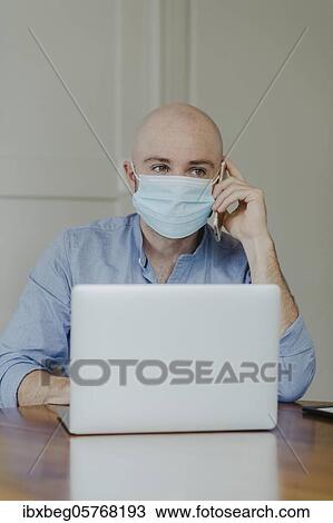 Stock Image - Man with face mask at home office. Fotosearch