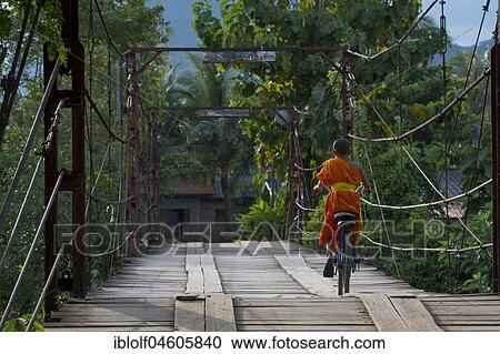 Stock Image - Novice, young monk, with bicycle on wooden bridge over the Nam Song River, Vang Vieng, Laos, Asia. Fotosearch