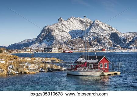 Stock Photo - Red wooden cabin on raft with sailing boat in the sea off the coast, behind fishing village and mountains, Seloy, Nordland, Norway, Europe. Fotosearch