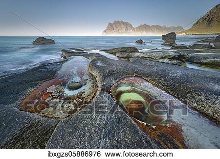 Stock Photograph - Rocks on the beach, in front tidal pond with formation Eye of Utakleiv, Nordland, Lofoten, Norway, Europe. Fotosearch