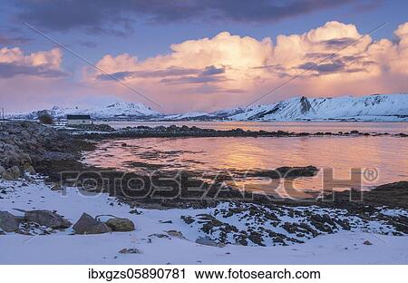 Stock Image - Snow-covered landscape with wooden house at sea bay and fjord, cloud atmosphere, warm evening light, Nordland, Lofoten, Norway, Europe. Fotosearch