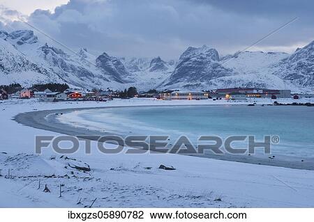 Stock Image - Snowy landscape at sea bay with sandy beach, behind village, dwellings and industrial buildings and high mountains, Ramberg, Nordland, Lofoten, Norway, Europe. Fotosearch
