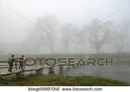 Stock Photograph - Two men walk in the morning fog over a bridge over the Rapti River, Sauraha, Chitwan National Park, Nepal, Asia. Fotosearch