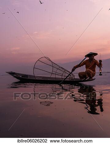 Stock Image - A traditional fisherman sits with a lamp on his small boat, at sunrise, Lake Inle, Myanmar, Asia. Fotosearch