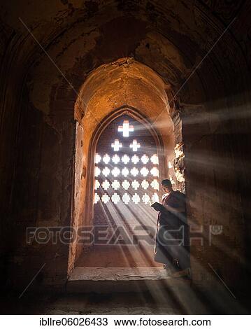 Stock Image - Buddhist young monk in red robe reading standing in front of rays of light in a temple, Bagan, Myanmar, Asia. Fotosearch