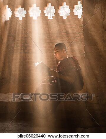 Stock Image - Buddhist young monk in red robe reading sitting in front of rays of light in a temple, Bagan, Myanmar, Asia. Fotosearch
