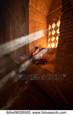 Stock Image - Buddhist young monk in red robe reading sitting in front of rays of light in a temple, Bagan, Myanmar, Asia. Fotosearch