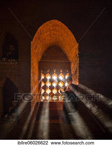 Stock Image - Buddhist young monk in red robe reading standing in front of rays of light in a temple, Bagan, Myanmar, Asia. Fotosearch