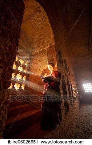 Stock Image - Buddhist young monk in red robe reading standing in front of rays of light in a temple, Bagan, Myanmar, Asia. Fotosearch