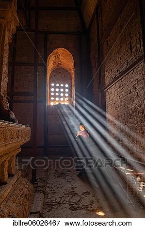 Stock Photo - Buddhist young monk in red robe reading standing in front of rays of light in a temple, Bagan, Myanmar, Asia. Fotosearch