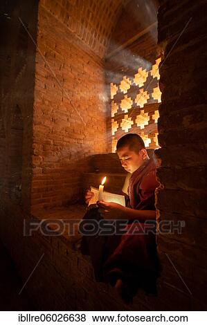 Stock Photo - Buddhist young monk in red robe reading while sitting with a candle in front of rays of light in a temple, Bagan, Myanmar, Asia. Fotosearch