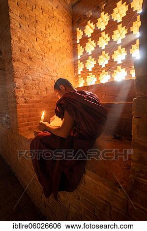 Buddhist young monk in red robe reading while sitting with a candle in front of rays of light in a temple, Bagan, Myanmar, Asia View Large Photo Image Stock Photograph - Buddhist young monk in red robe reading while sitting with a candle in front of rays of light in a temple, Bagan, Myanmar, Asia. Fotosearch