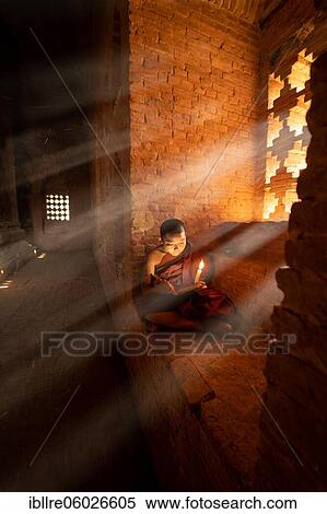 Stock Photography - Buddhist young monk in red robe sits and reads rays of light in a temple, Bagan, Myanmar, Asia. Fotosearch